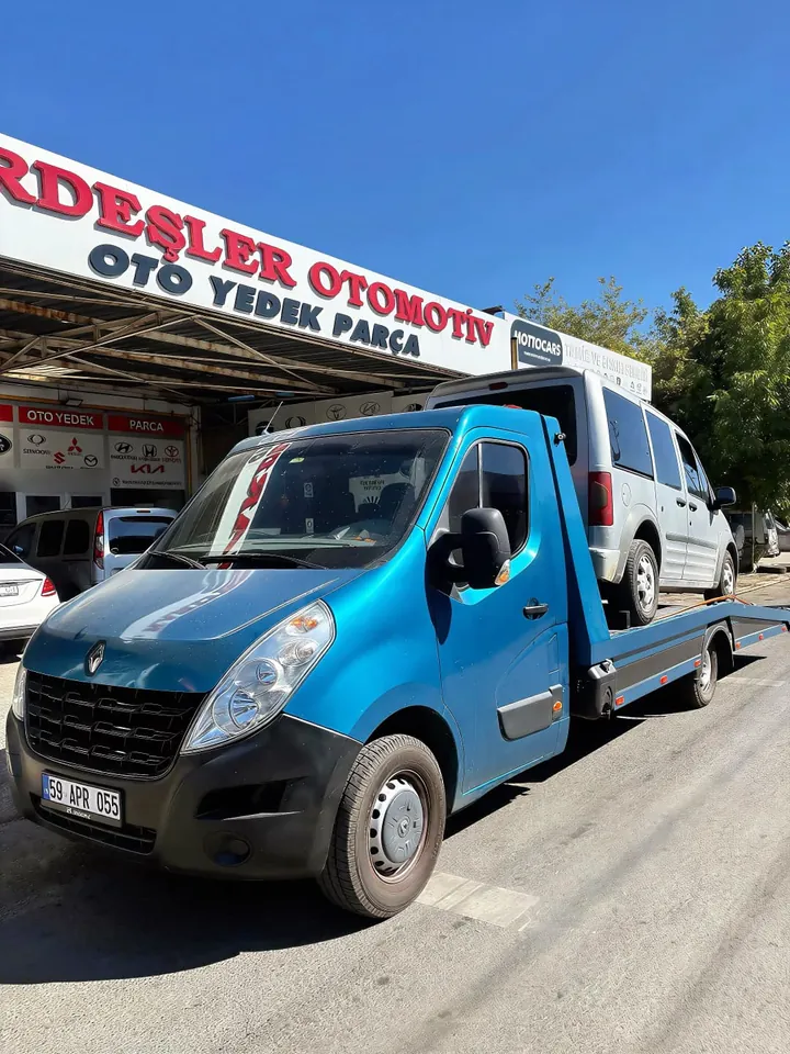 Platformlu çekici, gri hafif ticari aracı oto yedek parça dükkânı önünde taşıyor